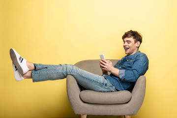 cheerful young man sitting in armchair and using smartphone on yellow background