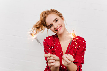 Pleasant young lady in red attire holding bengal lights. Indoor photo of excited girl with wavy...