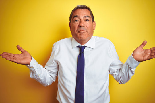 Handsome middle age businessman standing over isolated yellow background clueless and confused expression with arms and hands raised. Doubt concept.