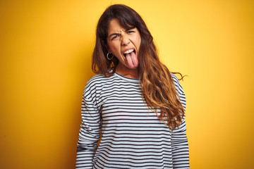 Young beautiful woman wearing stripes t-shirt standing over yelllow isolated background sticking tongue out happy with funny expression. Emotion concept. © Krakenimages.com