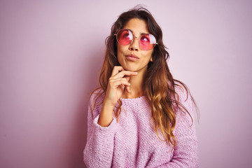 Young beautiful woman wearing fashion sunglasses standing over pink isolated background with hand on chin thinking about question, pensive expression. Smiling with thoughtful face. Doubt concept.