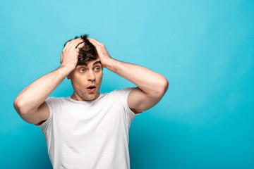 shocked young man looking away while holding hands on head on blue background