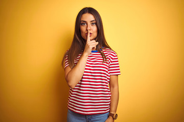 Young beautiful woman wearing striped t-shirt standing over isolated yellow background asking to be quiet with finger on lips. Silence and secret concept.