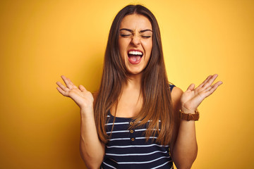 Young beautiful woman wearing striped t-shirt standing over isolated yellow background celebrating mad and crazy for success with arms raised and closed eyes screaming excited. Winner concept