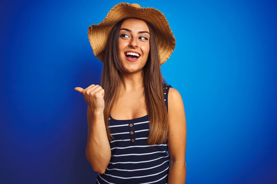 Young beautiful woman wearing striped t-shirt and summer hat over isolated blue background smiling with happy face looking and pointing to the side with thumb up.