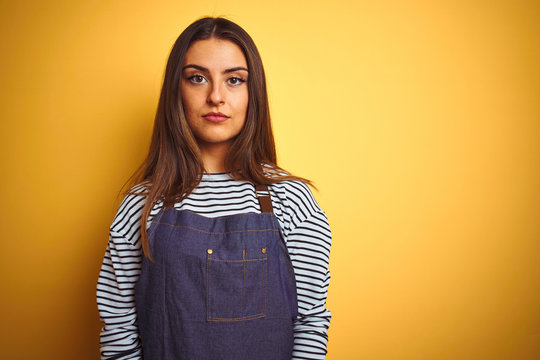 Young beautiful baker woman wearing apron standing over isolated yellow background with serious expression on face. Simple and natural looking at the camera.