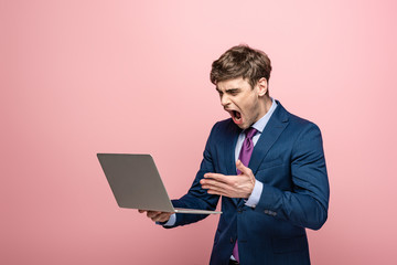 angry businessman quarreling while looking at laptop isolated on pink