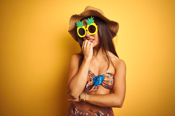 Woman on vacation wearing bikini and pineapple sunglasses over isolated yellow background looking stressed and nervous with hands on mouth biting nails. Anxiety problem.