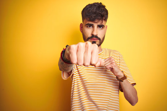 Young man with tattoo wearing striped t-shirt standing over isolated yellow background Punching fist to fight, aggressive and angry attack, threat and violence