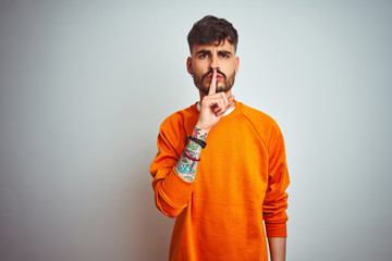 Young man with tattoo wearing orange sweater standing over isolated white background asking to be quiet with finger on lips. Silence and secret concept.