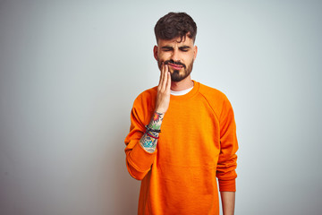 Young man with tattoo wearing orange sweater standing over isolated white background touching mouth...