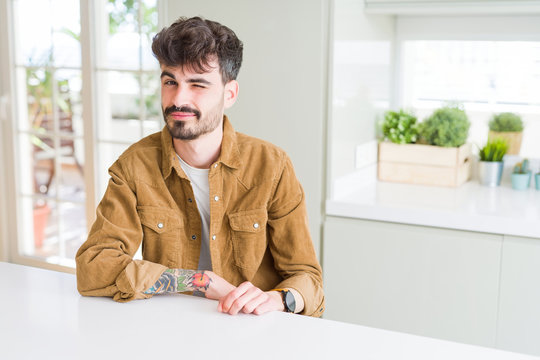 Young man wearing casual jacket sitting on white table winking looking at the camera with sexy expression, cheerful and happy face.