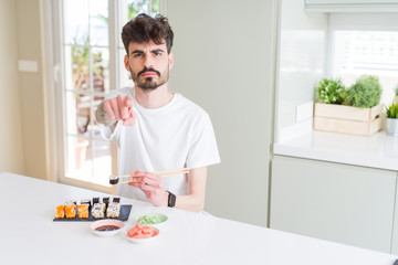 Young man eating asian sushi from home delivery pointing with finger to the camera and to you, hand sign, positive and confident gesture from the front