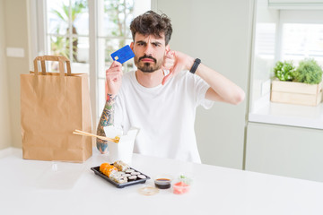 Young man eating asian sushi from home delivery using credit card as payment with angry face, negative sign showing dislike with thumbs down, rejection concept
