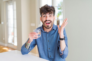 Young man drinking a fresh glass of water very happy and excited, winner expression celebrating victory screaming with big smile and raised hands