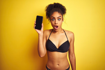 African american woman wearing bikini showing smartphone over isolated yellow background scared in...