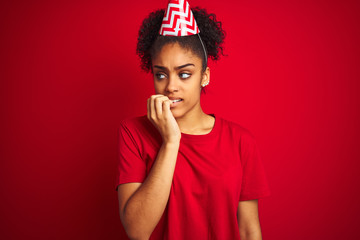 Young afro american woman wearing birthday hat over isolated red background looking stressed and nervous with hands on mouth biting nails. Anxiety problem.