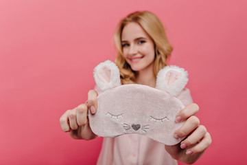 Indoor portrait of chilling blonde girl with cute eyemask on foreground. Blur photo of joyful fair-haired female model with pink sleep mask in focus.
