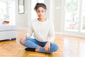 Beautiful young african american woman with afro hair sitting on the floor with serious expression on face. Simple and natural looking at the camera.