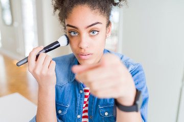 Young african american girl appying blush using make up brush pointing with finger to the camera and to you, hand sign, positive and confident gesture from the front