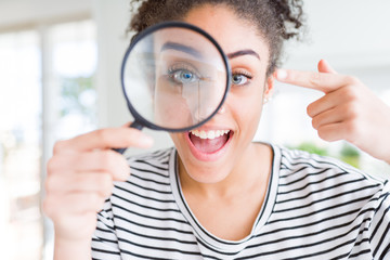 Young african american woman looking through magnifying glass very happy pointing with hand and...