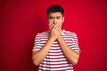 Young asian chinese man wearing striped t-shirt standing over isolated red background shocked covering mouth with hands for mistake. Secret concept.