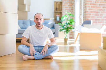 Young bald man sitting on the floor around cardboard boxes moving to a new home afraid and shocked with surprise expression, fear and excited face.