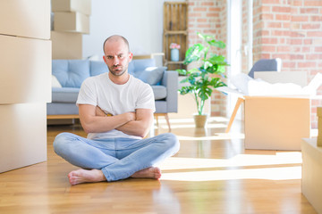 Young bald man sitting on the floor around cardboard boxes moving to a new home skeptic and nervous, disapproving expression on face with crossed arms. Negative person.