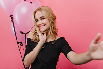 Happy fair-haired woman in black clothes making selfie on pink background. Glad birthday girl having fun with helium balloons.