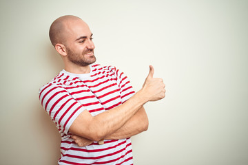 Fototapeta premium Young bald man with beard wearing casual striped red t-shirt over white isolated background Looking proud, smiling doing thumbs up gesture to the side