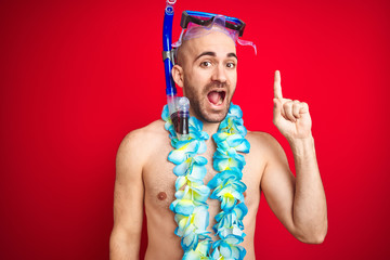 Young man wearing diving snorkel goggles and hawaiian lei flowers over isolated red background...