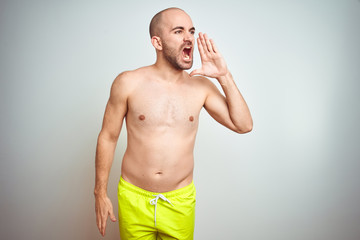 Young shirtless man on vacation wearing yellow swimwear over isolated background shouting and screaming loud to side with hand on mouth. Communication concept.