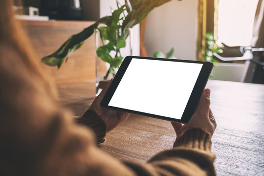 Mockup image of hands holding and using black tablet pc with blank white desktop screen on wooden table