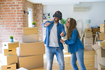Young couple moving to a new house having fun playing with virtual reality glasses around cardboard boxes