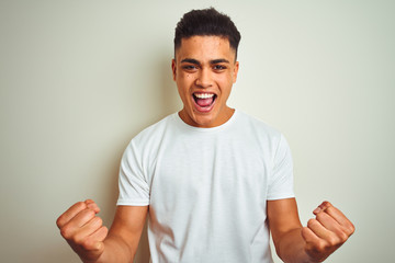 Young brazilian man wearing t-shirt standing over isolated white background celebrating surprised and amazed for success with arms raised and open eyes. Winner concept.