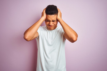 Young brazilian man wearing t-shirt standing over isolated pink background suffering from headache desperate and stressed because pain and migraine. Hands on head.