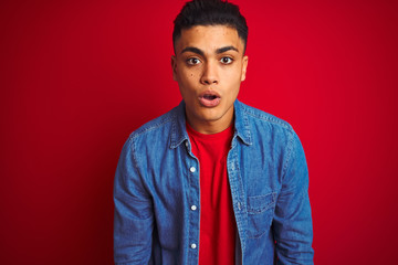 Young brazilian man wearing denim shirt standing over isolated red background afraid and shocked with surprise expression, fear and excited face.