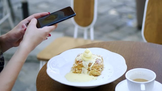 Woman in street cafe making picture of beautiful appetising cake with white creem on her phone