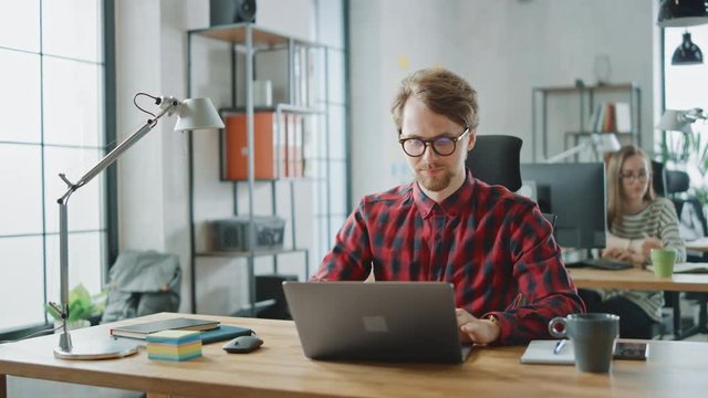 Handsome Young Man in Glasses and Shirt is Working on a Laptop in a Creative Business Agency. They Work in Loft Office. Diverse People Working in the Background. He's in Good Mood.