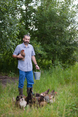 A smiling cute bearded farmer holds a chicken in one hand and in the other a metal bucket, the young man stands on a blurred background of green trees