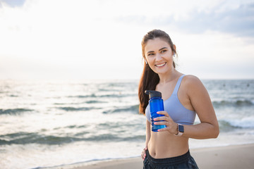 Young brunette woman with wireless earbuds and smart watches resting after morning workout at the sea shore at sunrise drinking water