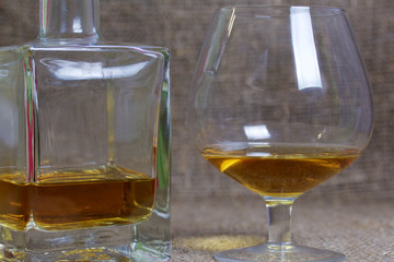 A bottle of strong alcohol. From transparent glass, a square form with a golden stopper. Near a glass with a drink poured. Against the backdrop of coarse linen fabric. Close-up.