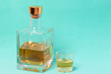 A bottle of strong alcohol. From transparent glass, a square form with a golden stopper. Near a stack with a drink poured. Against the background of mint color.