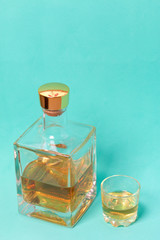 A bottle of strong alcohol. From transparent glass, a square form with a golden stopper. Near a stack with a drink poured. Against the background of mint color.