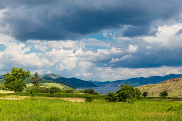 Fototapeta premium Blue sky with white clouds, trees, fields and meadows with green grass, against the mountains. Composition of nature. Rural summer landscape.