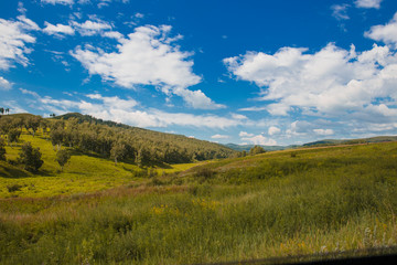 Blue sky with white clouds, trees, fields and meadows with green grass, against the mountains. Composition of nature. Rural summer landscape.