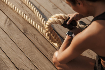 Cropped image of focused young woman looking at wristwatch while doing workout with battle ropes