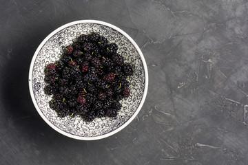 Fresh raw black mulberries in plate on dark concrete background. Top view with copy space. Flat lay background