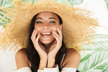 Photo closeup of seductive brunette woman in straw hat and beach clothes smiling at camera