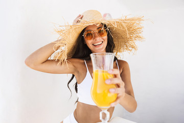 Photo of cheerful brunette woman in straw hat and swimsuit smiling while standing over white wall...